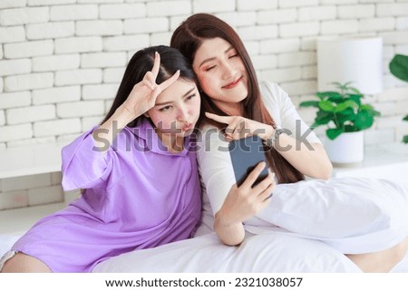 Two Asian cheerful happy female girlfriends in casual outfits sitting resting relaxing holding touchscreen smartphone showing two finger sign smiling taking selfie photo together on bed in bedroom.