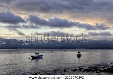 the charm and beauty of clouds with mountains covered in mist, and fishing boats anchored near the beach with calm sea conditions at sunset