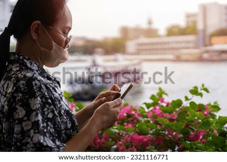 Side view of Asian woman is using a smartphone on the bank of the river, along which a passenger ship is traveling. people and technology.