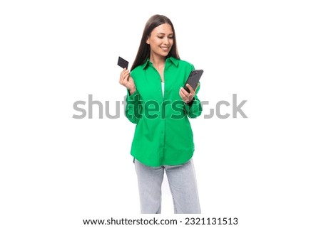 cute young brown-haired female model with brown eyes in a green shirt pays for purchases online using a mobile phone and a card