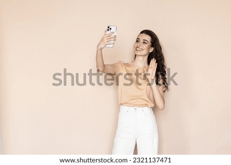 young woman on a beige background waving her hand at the phone taking a selfie or talking via video call