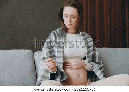Young ill pregnant woman wears casual clothes plaid hold thermometer sits on grey sofa couch stay at home hotel flat rest relax spend free spare time in living room indoor. Maternity family concept