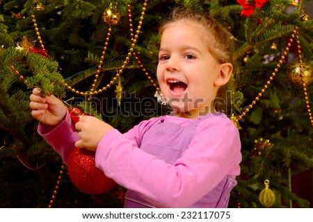 Adorable toddler girl holding decorative toy ball on Christmas tree branch