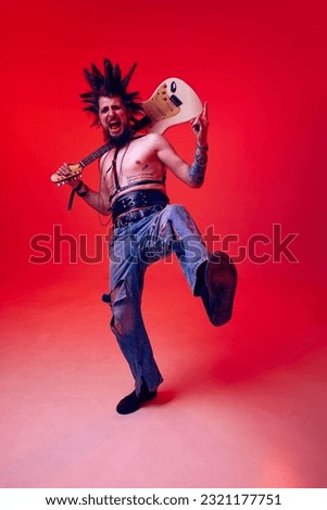 Portrait of creative young man, punk, musician posing with electric guitar against red studio background in neon light. Concept of music, lifestyle, subculture, art, youth, human emotions