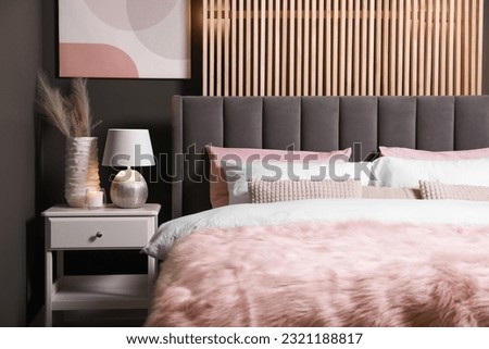Comfortable bed with cushions, lamp and different decor on white bedside table in room. Stylish interior