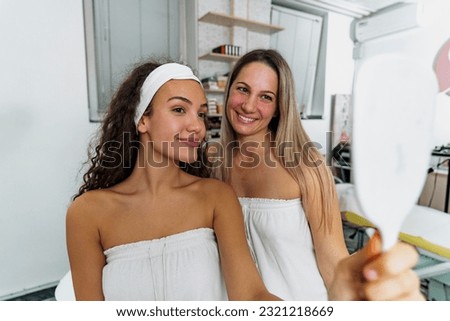 Picture of a two young woman wrapped in a white towel looking at themselves in a mirror after a spa treatment