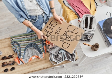 top view of cropped tattooed woman holding swap not shop card near sunglasses, clothes, electric toaster, vinyl record player and cezve, sustainable living and circular economy concept Royalty-Free Stock Photo #2321232859