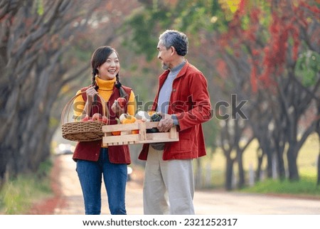 a daughter holding basket of fruits and father carrying wooden crate fill with pumpkins smile and talk about quality of agricultural productivity while walking along the street at farmland in autumn