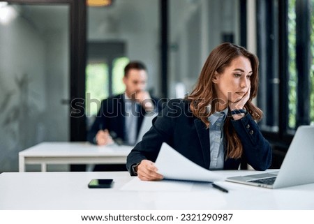A hardworking female employee wearing a suit, sitting at the office table, working on a laptop while her male colleague working behind her. Royalty-Free Stock Photo #2321290987