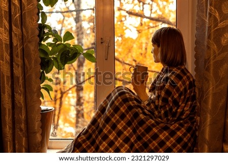 Autumn coffee. A beautiful girl with a cup of coffee in her hands sits and looks out the window. Cozy autumn composition. Copy space. Royalty-Free Stock Photo #2321297029