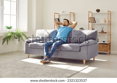 Young relaxed man sitting on comfortable couch in the living room at modern home and having a rest from a work with closed eyes. Calm peaceful guy enjoying quiet time alone in the room and dreaming. Royalty-Free Stock Photo #2321323537