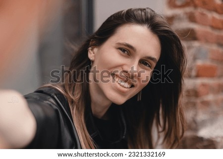 Pensive brunette woman make selfie posing on cafe outside background. Outdoor shot of happy hippie lady make video call. Girl wear black leather shirt, earrings raises her hand to camera.