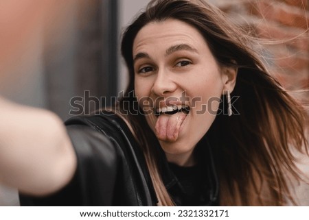 Pensive brunette woman make selfie posing on cafe outside background. Outdoor shot of happy hippie lady make video call. Girl wear black leather shirt, earrings raises her hand to camera, show tongue.