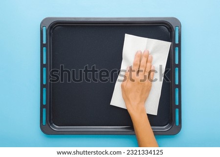 Young adult woman hand wiping dark black oven tray with dry white paper napkin on light blue table background. Pastel color. Closeup. Point of view shot. Royalty-Free Stock Photo #2321334125