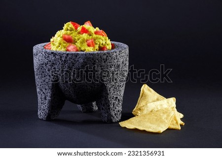 Delicious avocado guacamole with red diced tomatoes in a typical stone Mexican mortar or molcajete by a few totopos or nachos over a black background. Royalty-Free Stock Photo #2321356931