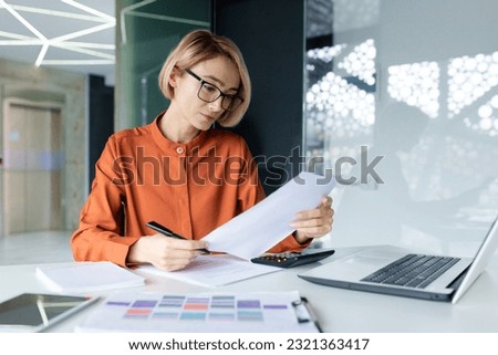 Serious and pensive business woman behind paper work inside office, female financier worker thinks about contracts and reports with charts and graphs, blonde successful woman uses laptop at work. Royalty-Free Stock Photo #2321363417