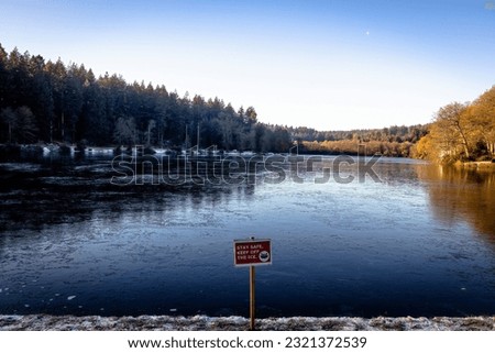 A "stay safe, keep off the ice" sign by the lake surrounded by trees under blue sky in winter