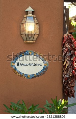 A vertical closeup a "welcome" sign in Spanish on a wall next to a lamp and dried chilies in Santa Fe