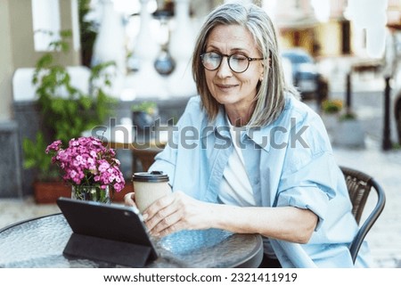 Mature lady enjoying outdoors while searching for information on tablet PC. Woman seated at cafe table on street, sipping cup of coffee. Moment of leisure and relaxation. High quality photo