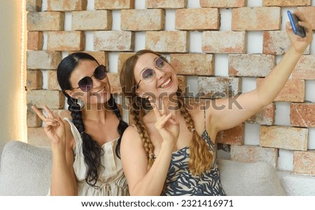 Ukrainian woman (brunette) and Russian woman (blonde) portrait together with braids posing and taking a selfie, showing peace gesture