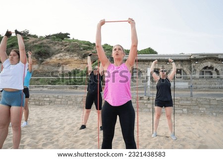 Women of various ages doing fitness workouts in class exercise with coach on the beach. Ladies working with resistance bands. Sport for health and wellbeing. Active lifestyle. Selective focus.