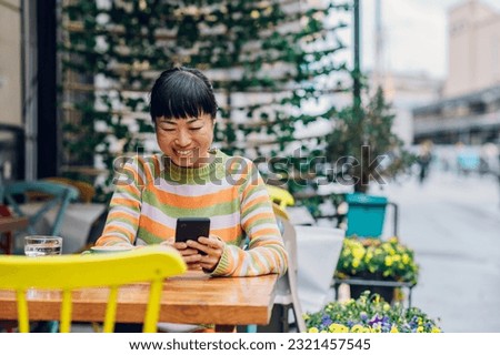 Cropped shot of a beautiful joyful asian woman sitting in a cozy outdoor restaurant while looking down to her phone with a big smile on her face. Enjoying a pleasant atmosphere while working remote.