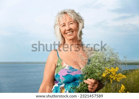 Smiling elderly woman in a summer sundress holding flowers in a field. Love for nature and relaxation. Royalty-Free Stock Photo #2321457589