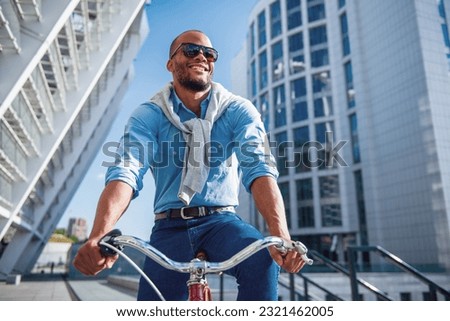 Low angle view of handsome young businessman in casual clothes and sun glasses riding his bike and smiling Royalty-Free Stock Photo #2321462005
