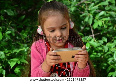 little girl with a phone and headphones outdoors in a garden or park. watching cartoons, playing games. modern kids and gadgets. Smiling girl listening music using smartphone wearing white headphones.