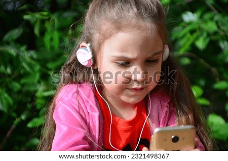 little girl with a phone and headphones outdoors in a garden or park. watching cartoons, playing games. modern kids and gadgets. Smiling girl listening music using smartphone wearing white headphones.