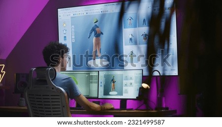 Young 3D designer creates video game character or clothes, works remotely from home in the evening on computer and big digital screen with professional software interface for 3D modeling and design.