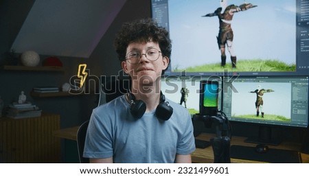 Young 3D designer speaks on camera using microphone, records video tutorial for blog about video game design and 3D modeling at home studio. Software interface on computer and big digital screen.