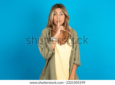 Surprised young beautiful blonde woman wearing overshirt makes silence gesture, keeps finger over lips and looks mysterious at camera