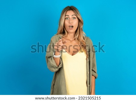 Shocked young beautiful blonde woman wearing overshirt points at you with stunned expression