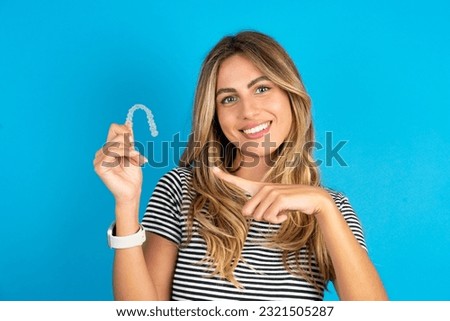 Young beautiful woman wearing striped t-shirt holding an invisible aligner and pointing at it. Dental healthcare and confidence concept.