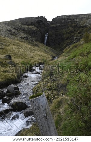 Stream flowing between sloping hills past a fence post from a waterfall slicing through a rock cliff