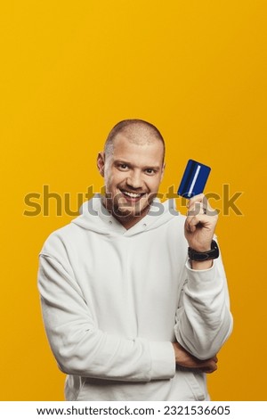 Vertical photo of smiling handsome guy holding credit card while smiling and looking at camera over yellow background, wearing white hoodie