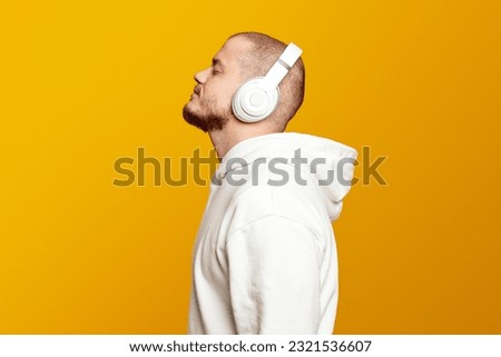 Side view of calm caucasian man wearing white hoodie and headphones, listening music with closed eyes, isolated over yellow background Royalty-Free Stock Photo #2321536607