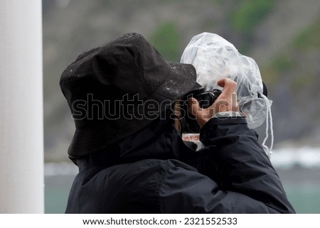 a women wearing a black rain hat and black rain coat holds a camera covered in plastic to her eye to take a photo in the rain Royalty-Free Stock Photo #2321552533