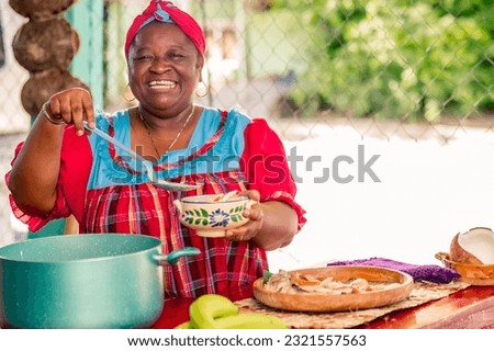 Beautiful black woman of the Garífuna ethnic group smiles at the camera as she puts the hot soup on the plate. Royalty-Free Stock Photo #2321557563