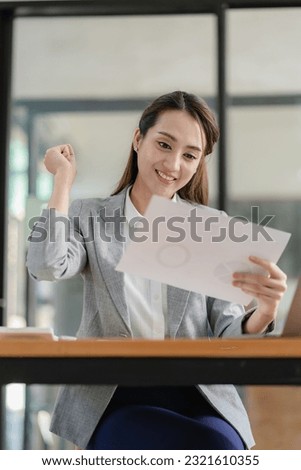Happy young asian businesswoman celebrating success with arms raised in front of laptop with excitement at office