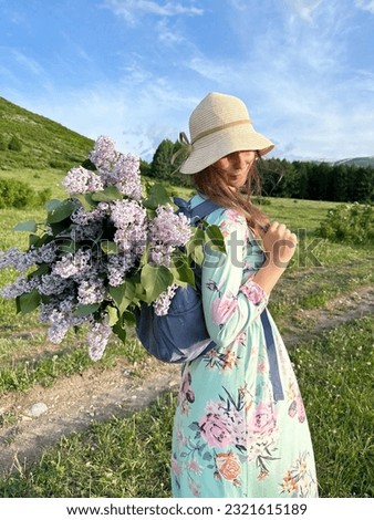 a girl in a dress and hat with a backpack and a bouquet of lilacs walks in a meadow