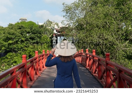 Asian woman is wearing Ao Dai traditional Vietnamese dress and traveling at Red Bridge- The Huc Bridge in Hoan Kiem Lake, this is a lake in the historical center of Hanoi, the capital city of Vietnam Royalty-Free Stock Photo #2321645253