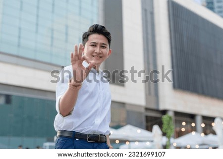 A handsome and confident asian man in his early 20s makes an okay sign. Outdoor city plaza scene.