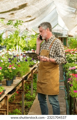 A vertical photograph capturing a dedicated floral nursery worker engaged in a phone conversation, jotting down a customer's flower order in a notebook.