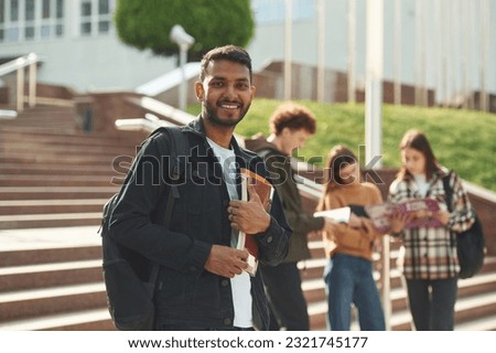 Indian man is standing in front of his friends. Four young students in casual clothes are together outdoors.