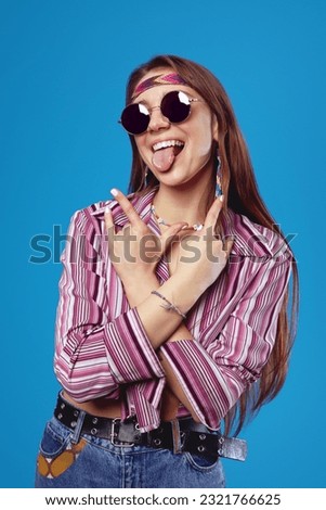 Excited hippie girl showing tongue and rock gesture with both hands crossed, dressed in pink shirt and round sunglasses, isolated on blue background