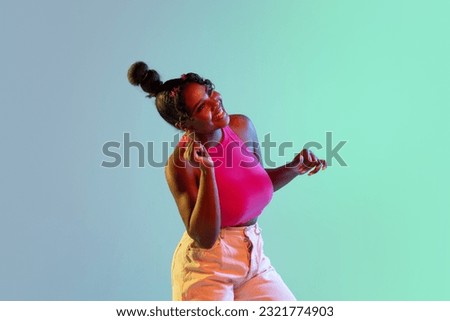 Joyful young African girl, female fashion model dancing at camera isolated on blue green background in neon light. Concept of human emotion, facial expression. Copy space for ad.