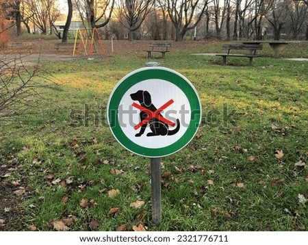 A "dogs prohibited" sign in a green circle on a pole in an outdoor park with benches and a swing in the background
