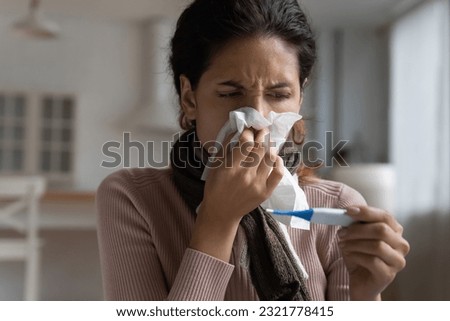 Unhealthy young latin woman wiping runny nose with paper tissue, measuring body temperature with electronic device. Worrying millennial hispanic lady having first flue grippe corona virus symptoms. Royalty-Free Stock Photo #2321778415